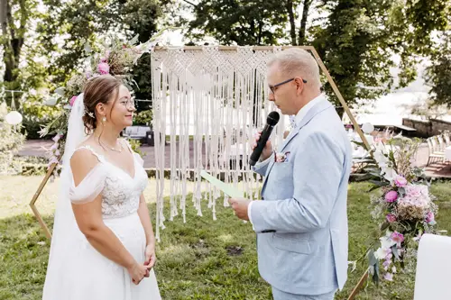 Eine Braut in weißem Kleid und Brille lächelt, während sie dem Bräutigam zuhört, der ein Mikrofon hält und aus einem grünen Blatt vorliest. Ein Hochzeitsfotograf in Marcolinis Welt hat die beiden während ihrer Hochzeitszeremonie im Freien mit Blumenschmuck und viel Grün festgehalten.