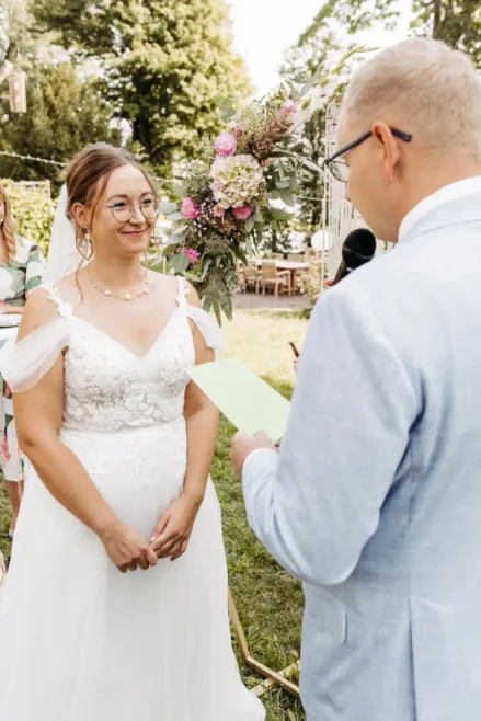 Eine Braut in weißem Kleid und Brille lächelt, während sie dem Bräutigam zuhört, der ein Mikrofon hält und aus einem grünen Blatt vorliest. Ein Hochzeitsfotograf in Marcolinis Welt hat die beiden während ihrer Hochzeitszeremonie im Freien mit Blumenschmuck und viel Grün festgehalten.