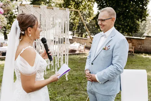 Eine Braut im weißen Kleid, mit Mikrofon in der Hand, liest ihrem Bräutigam im hellblauen Anzug ihre Eheversprechen vor. Sie stehen im Freien vor einem Makramee-Hintergrund, umgeben von üppigem Grün und sanftem Sonnenlicht – wunderschön eingefangen vom Hochzeitsfotografen in Marcolinis Welt.