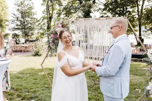 Ein lächelndes Paar hält Händchen während einer Zeremonie im Freien, festgehalten von einem Hochzeitsfotografen in Marcolinis Welt. Die Frau trägt ein weißes Spitzenkleid und einen Blumenkopfschmuck, während der Mann einen hellblauen Anzug und eine Brille vor einem Makramee-Hintergrund und üppigem Grün trägt.