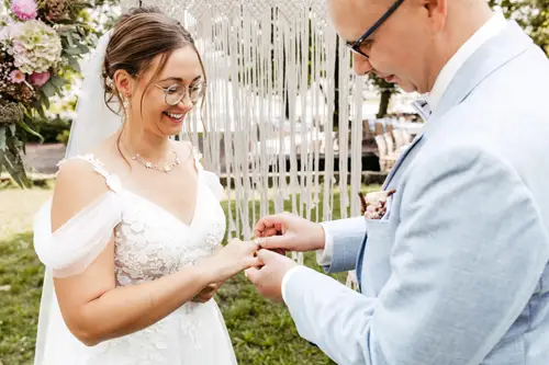 Während einer bezaubernden Zeremonie im Freien, die von einem Hochzeitsfotografen in Marcolinis Welt festgehalten wurde, tauschen Braut und Bräutigam die Ringe. Die Braut glänzt in einem weißen Spitzenkleid, während der Bräutigam in einem hellblauen Anzug im romantischen Licht der Lichterketten umwerfend wirkt.