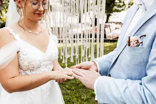Während einer bezaubernden Zeremonie im Freien, die von einem Hochzeitsfotografen in Marcolinis Welt festgehalten wurde, tauschen Braut und Bräutigam die Ringe. Die Braut glänzt in einem weißen Spitzenkleid, während der Bräutigam in einem hellblauen Anzug im romantischen Licht der Lichterketten umwerfend wirkt.