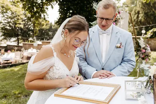 Eine Braut unterschreibt ein Dokument an einem Tisch im Freien, umgeben von üppigen Bäumen und elegantem Ambiente, während ihr Bräutigam im hellblauen Anzug zusieht. Ihr weißes Brautkleid und ihre Brille verleihen der Szene Charme, perfekt eingefangen von einem Hochzeitsfotografen in Marcolinis Welt.