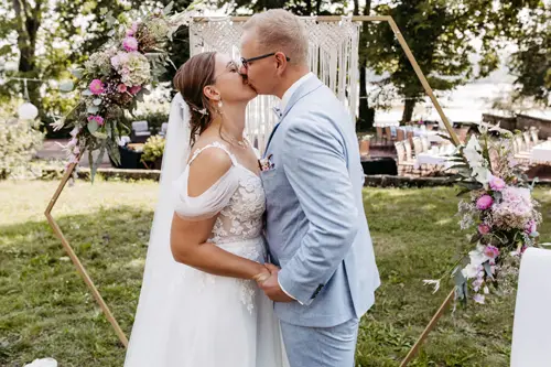 Braut und Bräutigam küssen sich unter einem atemberaubenden Blumenbogen, wunderschön eingefangen von Melanie Kunert Fotografie. Die Braut glänzt in ihrem schulterfreien Spitzenkleid, während der Bräutigam einen hellblauen Anzug trägt. Bäume rahmen diese bezaubernde Szene von Hochzeitsfotograf Dresden ein.