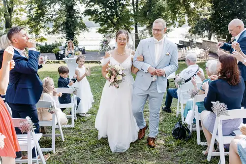 Braut und Bräutigam schreiten durch einen Gang im Freien, gesäumt von Gästen auf weißen Stühlen. Die Braut hält einen Blumenstrauß in ihrem weißen Kleid, während der Bräutigam einen hellgrauen Anzug trägt. Die Gäste klatschen und lächeln im Schatten der Bäume. Ihre Freude wurde von einem Hochzeitsfotografen in Marcolinis Welt eingefangen.