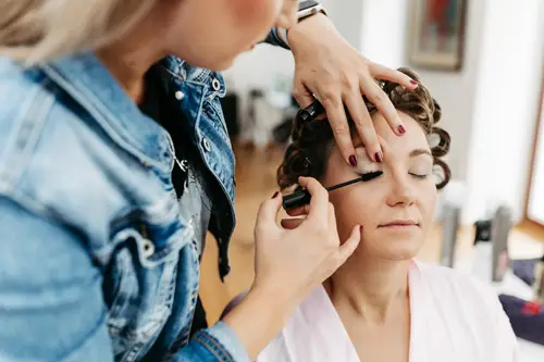 In einem Salon im Innenbereich, entspannt sich eine gelassene Person mit Lockenwicklern, während ihr sorgfältig Augen-Make-up aufgetragen wird und ein Moment festgehalten wird.