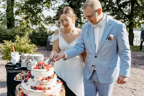 Braut und Bräutigam schneiden eine dreistöckige Hochzeitstorte an, die mit roten und schwarzen Beeren verziert ist. Ein Hochzeitsfotograf in Marcolinis Welt hat die Torte auf einem Holzständer, umgeben von verschiedenen Gebäckstücken, vor einer eleganten Außenkulisse mit Tischen und Stühlen festgehalten.
