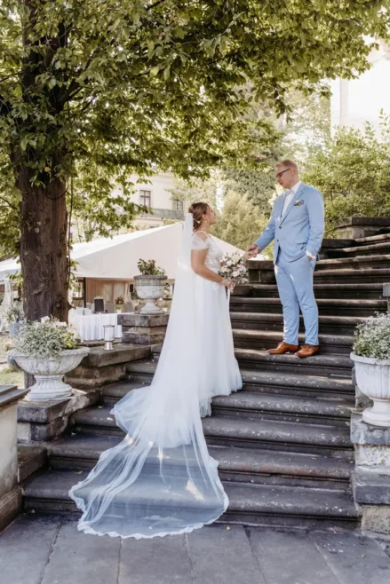 Eine Braut in einem fließenden weißen Kleid steht mit einem Blumenstrauß in der Hand auf einer Steintreppe und tauscht mit dem Bräutigam im hellblauen Anzug das Eheversprechen aus. Umgeben von üppigem Grün und Blumentöpfen wurde diese ruhige Szenerie im Freien vom Hochzeitsfotografen in Marcolinis Welt perfekt eingefangen.