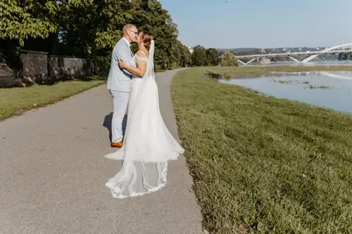Ein Paar in Hochzeitskleidung umarmt sich auf einem gepflasterten Weg neben einer Wiese und einem Fluss, wunderschön eingefangen von einem Hochzeitsfotografen in Marcolinis Welt. Der lange Schleier der Braut weht hinter ihr her, während sich im Hintergrund eine Brücke anmutig unter dem klaren blauen Himmel wölbt.