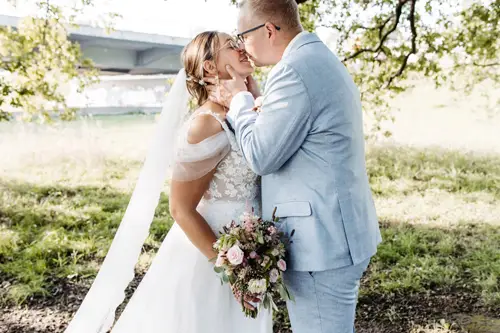 Ein talentierter Hochzeitsfotograf in Marcolinis Welt hält ein Paar fest, das sich an seinem Hochzeitstag liebevoll umarmt. Die Braut in einem floralen Spitzenkleid hält einen Strauß rosa und weißer Blumen in der Hand, während sie sich unter dem sonnendurchfluteten Baum herzlich anlächeln. Der Bräutigam trägt einen hellblauen Anzug.