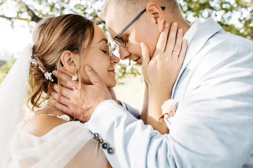 Ein talentierter Hochzeitsfotograf in Marcolinis Welt hält ein Paar fest, das sich an seinem Hochzeitstag liebevoll umarmt. Die Braut in einem floralen Spitzenkleid hält einen Strauß rosa und weißer Blumen in der Hand, während sie sich unter dem sonnendurchfluteten Baum herzlich anlächeln. Der Bräutigam trägt einen hellblauen Anzug.