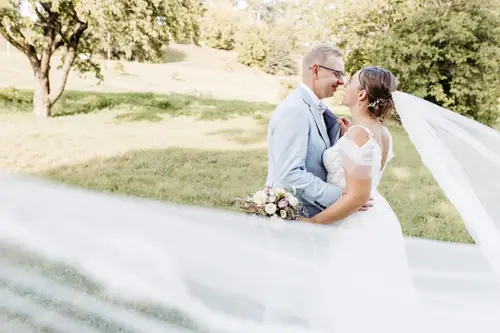 Braut und Bräutigam stehen auf einer Wiese, umarmt und lächelnd. Der Schleier der Braut weht im Wind, während Sonnenlicht durch eine große Eiche hinter ihnen fällt. Eingefangen von einem Hochzeitsfotografen in Marcolinis Welt, strahlt diese ruhige Szenerie Romantik aus.