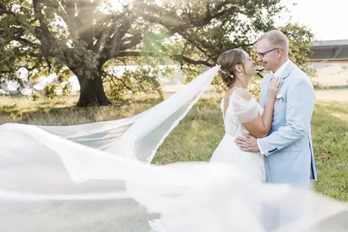Braut und Bräutigam stehen auf einer Wiese, umarmt und lächelnd. Der Schleier der Braut weht im Wind, während Sonnenlicht durch eine große Eiche hinter ihnen fällt. Eingefangen von einem Hochzeitsfotografen in Marcolinis Welt, strahlt diese ruhige Szenerie Romantik aus.