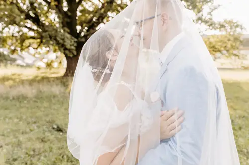 Braut und Bräutigam umarmen sich zärtlich unter einem Schleier, der Bräutigam legt sanft seine Stirn an die der Braut, während sie in elegantem Gewand dastehen. Eingefangen von einem talentierten Hochzeitsfotografen in Marcolinis Welt, unterstreichen ihre Brille und ihr Schleier diesen intimen Moment vor einem sanft verschwommenen Hintergrund.