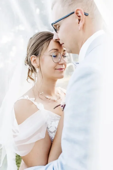 Braut und Bräutigam umarmen sich zärtlich, der Bräutigam legt sanft seine Stirn an die der Braut, während sie in elegantem Gewand dastehen. Eingefangen von einem talentierten Hochzeitsfotografen in Marcolinis Welt, unterstreichen ihre Brille und ihr Schleier diesen intimen Moment vor einem sanft verschwommenen Hintergrund.