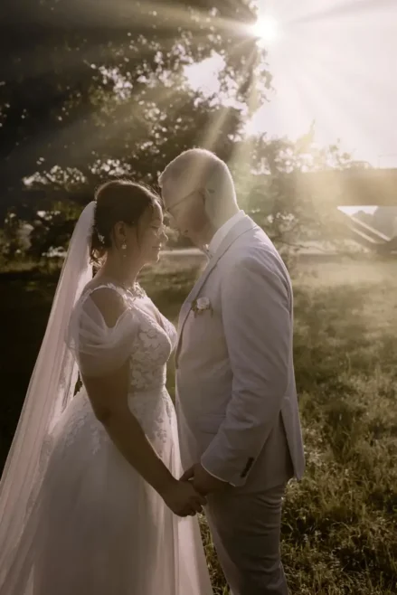 Ein Paar in Hochzeitskleidung steht dicht beieinander, die Köpfe berühren sich, lächelnd. Sonnenlicht zaubert einen warmen Schein hinter sie. Die Braut trägt ein Spitzenkleid und einen Schleier, während der Bräutigam einen hellgrauen Anzug mit Ansteckblume trägt. Ein Hochzeitsfotograf in Marcolinis Welt hält den perfekten Moment fest.