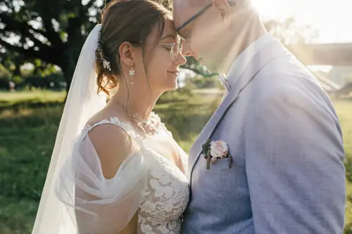 Ein Paar in Hochzeitskleidung steht dicht beieinander, die Köpfe berühren sich, lächelnd. Sonnenlicht zaubert einen warmen Schein hinter sie. Die Braut trägt ein Spitzenkleid und einen Schleier, während der Bräutigam einen hellgrauen Anzug mit Ansteckblume trägt. Ein Hochzeitsfotograf in Marcolinis Welt hält den perfekten Moment fest.