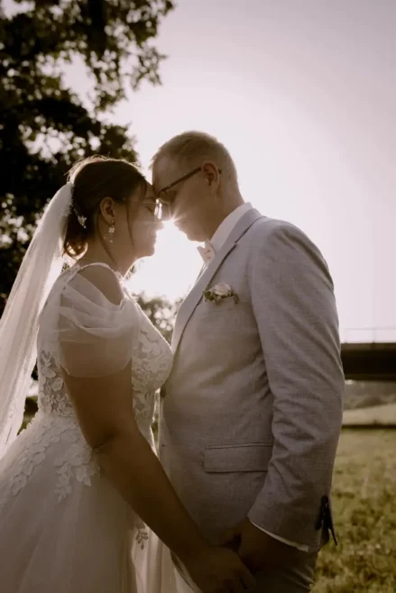 Ein Paar in Hochzeitskleidung steht dicht beieinander, die Köpfe berühren sich, lächelnd. Sonnenlicht zaubert einen warmen Schein hinter sie. Die Braut trägt ein Spitzenkleid und einen Schleier, während der Bräutigam einen hellgrauen Anzug mit Ansteckblume trägt. Ein Hochzeitsfotograf in Marcolinis Welt hält den perfekten Moment fest.