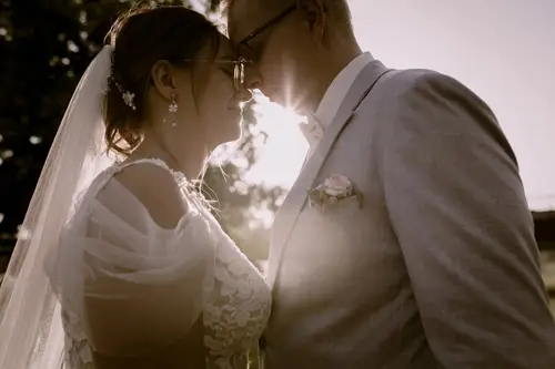Ein Paar in Hochzeitskleidung steht dicht beieinander, die Köpfe berühren sich, lächelnd. Sonnenlicht zaubert einen warmen Schein hinter sie. Die Braut trägt ein Spitzenkleid und einen Schleier, während der Bräutigam einen hellgrauen Anzug mit Ansteckblume trägt. Ein Hochzeitsfotograf in Marcolinis Welt hält den perfekten Moment fest.