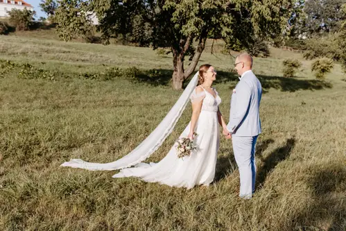 Vor der bezaubernden Kulisse von Marcolinis Welt hält eine Braut in einem weißen Kleid mit langem Schleier einen Blumenstrauß in der Hand und lächelt ihrem Bräutigam im hellblauen Anzug zu. Eingefangen von einem talentierten Hochzeitsfotografen, stehen sie Hand in Hand auf einer Wiese, eingerahmt von üppigen Bäumen.