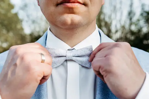 Nahaufnahme einer Person, die eine hellgraue Fliege zurechtrückt. Sie trägt einen hellblauen Anzug und ein weißes Hemd – die elegante Note wurde von einem Hochzeitsfotografen in Marcolinis Welt perfekt eingefangen. Ein goldener Ring ziert ihre linke Hand, während sanft verschwommenes Grün im Hintergrund den Charme des Augenblicks unterstreicht.