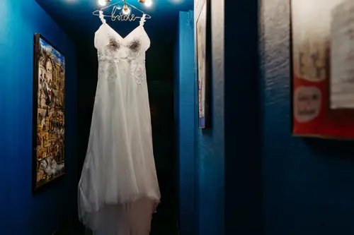 Ein weißes Brautkleid mit der Aufschrift „Braut“ hängt elegant in einem schmalen Flur mit leuchtend blauen Wänden, eingefangen von einem Hochzeitsfotografen in Marcolinis Welt. Das gedämpfte Licht und die gerahmten Bilder bilden einen künstlerischen Kontrast zu dieser bezaubernden Szene.