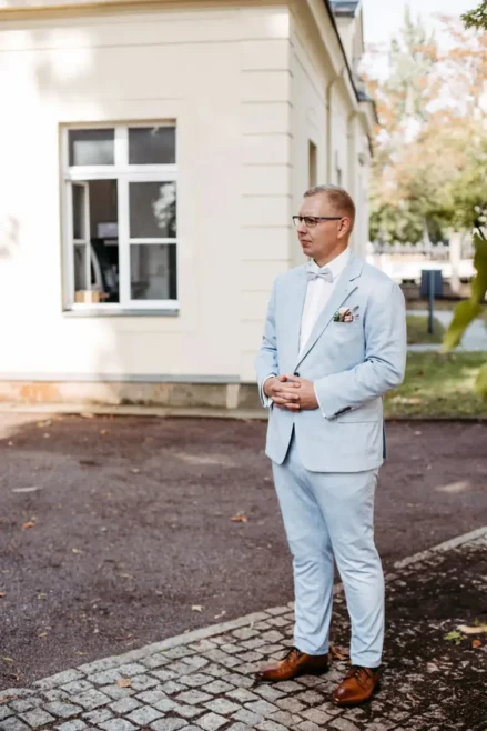Ein Mann im hellblauen Anzug, möglicherweise ein Hochzeitsfotograf in Marcolinis Welt, steht auf einem Kopfsteinpflasterweg in der Nähe eines hellen Gebäudes. Er trägt Brille, Fliege und braune Schuhe und verschränkt die Hände vor der Brust. Bäume und Blätter verleihen dem Hintergrund einen besonderen Charme.