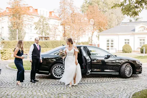 Eine Braut im weißen Kleid steigt anmutig mit dem Brautstrauß in der Hand aus einem schwarzen Auto. Begleitet von einem Mann in Weste und einer Frau im Overall schreiten die drei über die gepflasterte Auffahrt. Eingefangen von Hochzeitsfotograf Marcolinis Welt, wird dieser Moment von eleganten Bäumen und majestätischen Gebäuden eingerahmt.