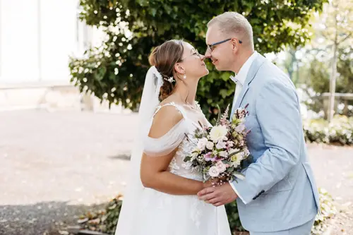 Eine Braut im weißen Kleid und ein Bräutigam im hellblauen Anzug teilen einen freudigen Moment. Nase an Nase stehen sie mit einem Blumenstrauß da. Ein Hochzeitsfotograf in Marcolinis Welt hielt sie fest, umgeben von üppigem Grün und in sanftes Sonnenlicht getaucht.