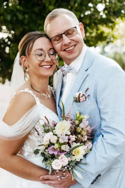 Ein glückliches Paar im Hochzeitskleid lächelt mit geschlossenen Augen. Die Braut hält einen Strauß weißer und rosa Blumen in der Hand und trägt ein Spitzenkleid mit Schleier. Der Bräutigam, in hellblauem Anzug und mit Brille, umarmt sie. Sie stehen im Freien, im Hintergrund eine grüne Landschaft.