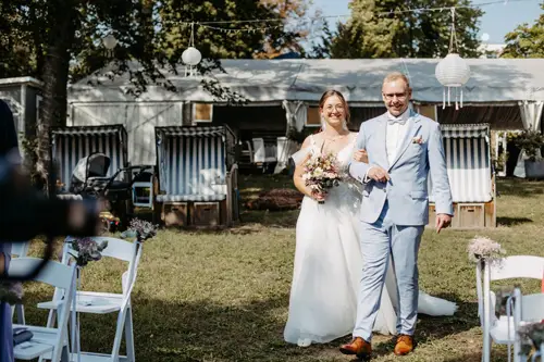 Braut und Bräutigam gehen lächelnd Arm in Arm einen Gartenweg entlang. Die Braut trägt ein weißes Kleid mit Blumenstrauß, der Bräutigam einen hellblauen Anzug. Weiße Stühle und Laternen schmücken die Rasenfläche, dahinter steht ein Zelt – wunderschön eingefangen von Melanie Kunert Fotografie, Hochzeitsfotografin Dresden.