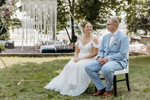 Braut und Bräutigam, gekleidet in ein weißes Spitzenbrautkleid und einen hellblauen Anzug, sitzen sich liebevoll anschaunend auf Stühlen. Der Anzug des Bräutigams ist mit einem Einstecktuch und einer Ansteckblume geschmückt. Diese ruhige Zeremonie im Freien auf einer Wiese wurde von Melanie Kunert Fotografie, der renommierten Hochzeitsfotografin Dresden, wunderschön eingefangen.