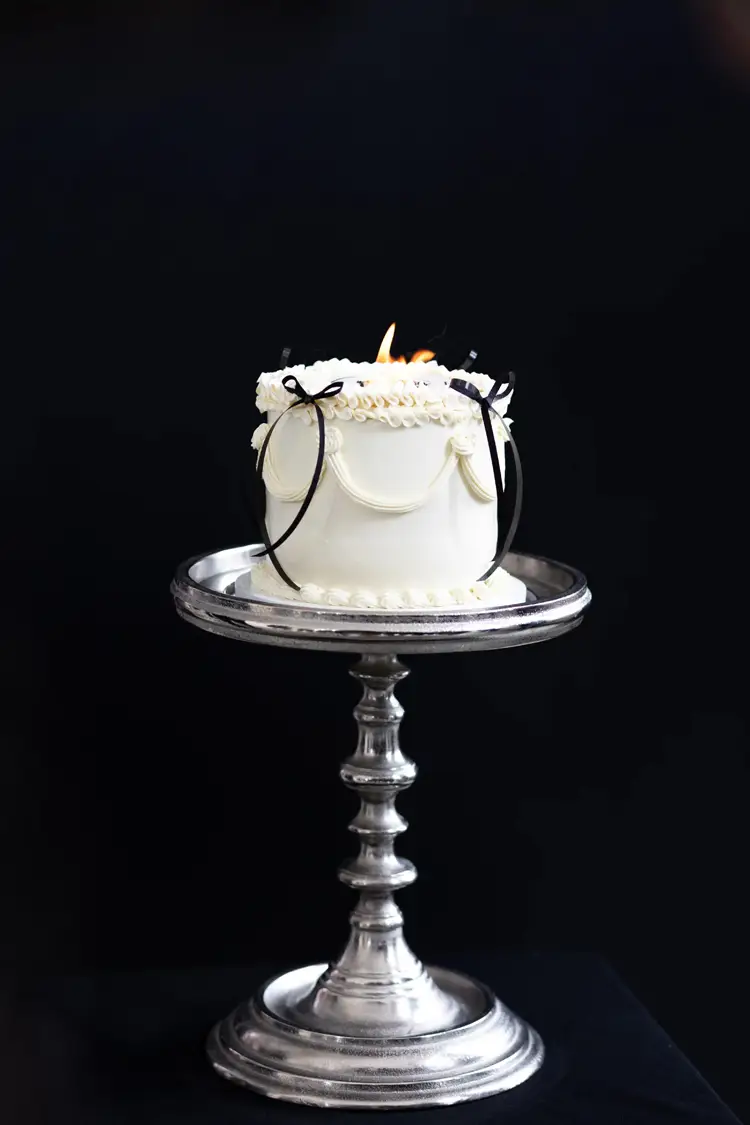 Eine kleine, runde weiße Torte mit kunstvollen Zuckerguss-Motiven steht auf einem verzierten silbernen Ständer. Schwarze Bänder zieren die Torte, in deren Mitte eine kleine Kerze flackert. Der schwarze Hintergrund des Modelshootings Solingen unterstreicht die elegante Optik der Torte.