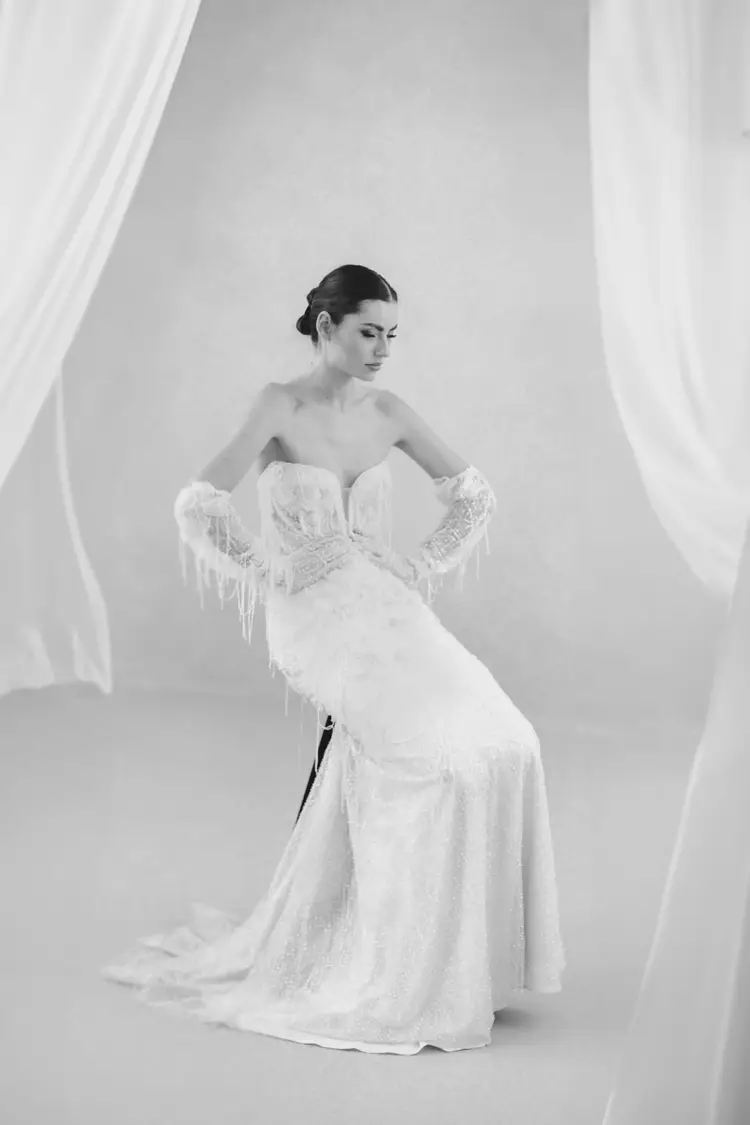 Eine Frau in einem trägerlosen, kunstvoll strukturierten weißen Kleid mit passenden langen Handschuhen sitzt anmutig während eines Modelshootings in Solingen. Die minimalistische Kulisse, eingerahmt von transparenten weißen Vorhängen, schafft eine sanfte und elegante Atmosphäre. Das Bild ist wunderschön in Schwarz-Weiß eingefangen.