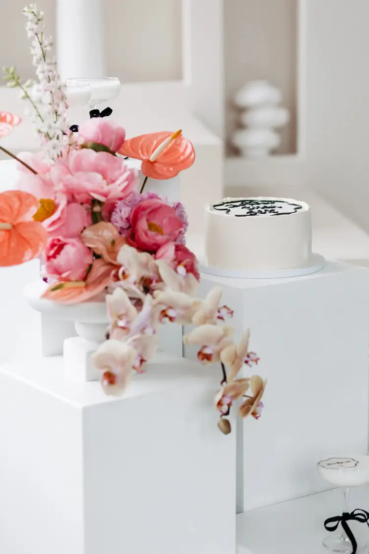 Eine weiße Torte mit schwarzer Zierpaspel steht auf einem weißen Podest, bereit für das Modelshooting Solingen. Links daneben ruht ein farbenfrohes Blumenarrangement mit rosa und orangefarbenen Blüten auf einem weißen Würfel. Der sanft verschwommene, neutrale Hintergrund unterstreicht diese elegante Präsentation.