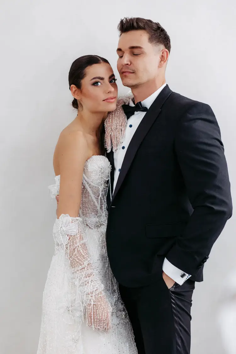 Ein Paar steht eng umschlungen bei einem Modelshooting in Solingen. Die Frau trägt ein elegantes weißes Spitzenbrautkleid mit Handschuhen, ihr Haar ist nach hinten frisiert. Der Mann trägt einen schwarzen Smoking mit Fliege und hat eine Hand in der Tasche. Beide wirken gelassen und zufrieden.
