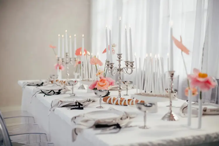Elegant gedeckter Esstisch für ein Event, perfekt für ein stilvolles Modelshooting in Solingen. Der Tisch ist mit hohen weißen Kerzen in silbernen Haltern, rosa Blumen und makellosem weißen Geschirr geschmückt. Ordentlich gefaltete Servietten und poliertes Silberbesteck runden das edle Ambiente ab.