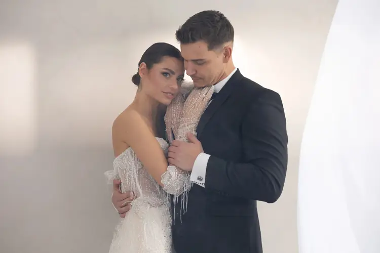 In einem sanft beleuchteten Raum, inmitten eines ruhigen Modelshootings in Solingen, umarmt sich ein Paar zärtlich. Die Frau im schulterfreien, perlenbesetzten weißen Kleid lehnt an dem Mann im schwarzen Anzug. Vor einem neutralen Hintergrund blicken sie sich liebevoll an.