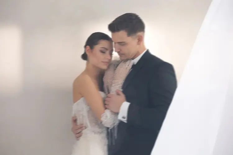 Bei einem Modelshooting in Solingen umarmt sich ein Paar zärtlich vor einem unscharfen Hintergrund. Die Frau in einem schulterfreien Spitzenbrautkleid legt ihren Kopf auf die Brust des Mannes. Der Mann, der einen dunklen Anzug trägt, legt seinen Arm um sie und schließt die Augen in heiterer Intimität.