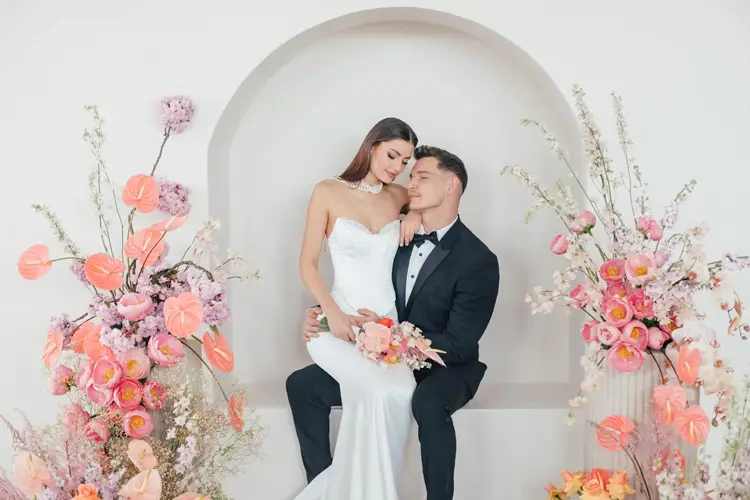 Eine Braut im weißen Kleid und ein Bräutigam im schwarzen Smoking sitzen in einer dekorativen Nische zusammen. Umgeben von rosa Blumen, die eine zauberhafte Kulisse für ihren intimen Moment schaffen, küsst der Bräutigam die Braut sanft auf die Stirn. Die perfekte Kulisse für ein Modelshooting in Solingen.