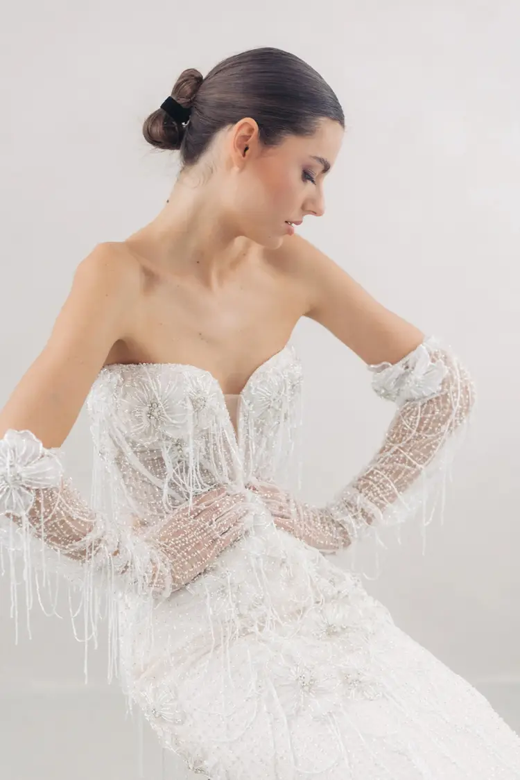 Bei einem Modelshooting in Solingen trägt eine Frau ein trägerloses weißes Kleid mit aufwendiger Perlenstickerei und dazu passende, ellenbogenlange Handschuhe. Ihr Haar ist zu einer glatten Hochsteckfrisur gestylt, während sie vor einem hellen Hintergrund seitlich nach unten blickt.