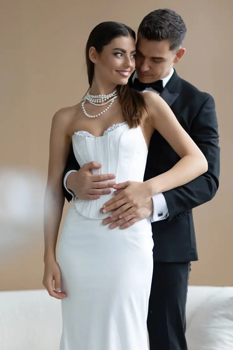 In einer ruhigen Umgebung, die an ein Modelshooting in Solingen erinnert, umarmt sich ein Paar liebevoll. Die Frau strahlt in ihrem weißen trägerlosen Kleid und der Perlenkette Eleganz aus, während der Mann sie in seinem schwarzen Smoking perfekt ergänzt. Beide lächeln herzlich vor einem neutralen Hintergrund im Innenbereich.