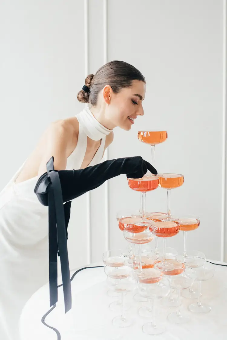 Eine Frau in weißem Kleid und schwarzen Handschuhen gießt während eines schicken Modelshootings in Solingen elegant Champagner in einen Turm aus Sektgläsern. Die Szene ist minimalistisch und vor einem hellen, neutralen Hintergrund gestaltet.