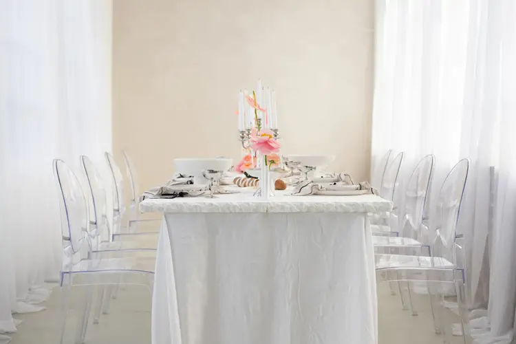 Ein minimalistisches Esszimmer-Setup, ideal für ein Modelshooting in Solingen, besteht aus einem langen, mit einer weißen Tischdecke bedeckten Tisch. Transparente Stühle säumen beide Seiten, während weiße Teller, Besteck und rosa Blumen ein elegantes Herzstück bilden. Weiche weiße Vorhänge hängen friedlich im Hintergrund.