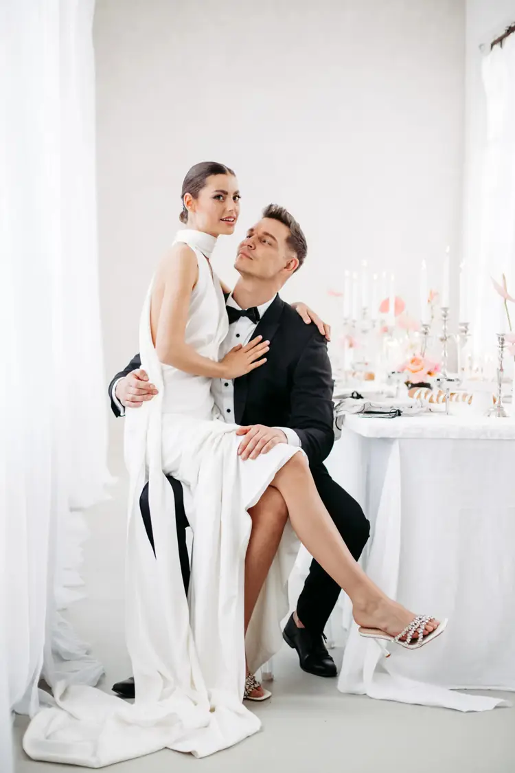 In einer edlen, hell erleuchteten Umgebung sitzt eine Frau in einem eleganten weißen Kleid anmutig auf dem Schoß eines Mannes – beide verkörpern Eleganz. Ihre Umgebung ist wunderschön mit Kerzen und Blumen geschmückt und erinnert an ein exklusives Modelshooting in Solingen.