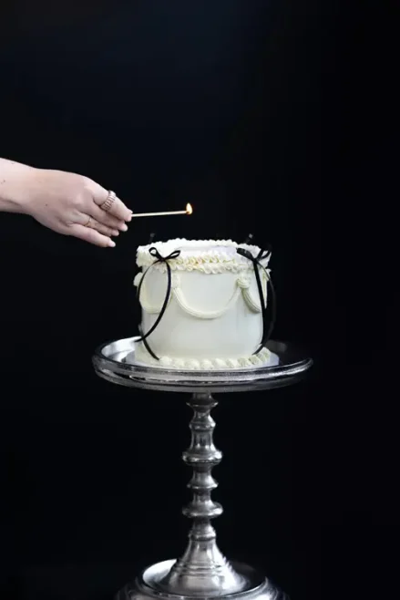 Eine Hand zündet eine Kerze auf einer weißen Torte an, die mit schwarzen Schleifen und dekorativem Zuckerguss verziert ist – perfekt für ein Model-Shooting in Solingen. Die Torte ruht elegant auf einem verzierten silbernen Ständer vor einem dramatischen schwarzen Hintergrund.