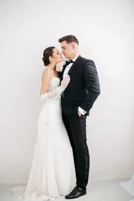 Bei einem Modelshooting in Solingen erleben Braut und Bräutigam einen intimen Moment. Die Braut, in einem detailreichen weißen Kleid mit Handschuhen, berührt sanft das Gesicht des Bräutigams. Der Bräutigam, im schwarzen Smoking, steht mit der Hand in der Tasche vor einem schlichten, hellen Hintergrund.