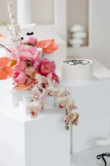 Eine weiße Torte mit schwarzer Zierpaspel steht auf einem weißen Podest, bereit für das Modelshooting Solingen. Links daneben ruht ein farbenfrohes Blumenarrangement mit rosa und orangefarbenen Blüten auf einem weißen Würfel. Der sanft verschwommene, neutrale Hintergrund unterstreicht diese elegante Präsentation.