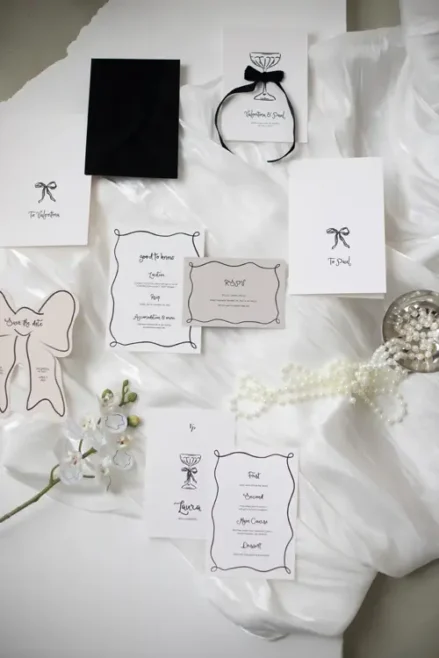 Elegante Hochzeitseinladungen und Papeterie sind auf weichem, weißem Stoff arrangiert und präsentieren schwarz-weiße Designs mit raffinierten Schleifen- und Ringmotiven. Verziert mit Perlen, einem schwarzen Umschlag und einem Hauch weißer Blumen fängt dieses Arrangement den Charme des Modelshootings Solingen ein.