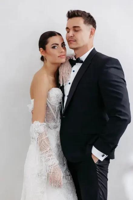 Ein Paar steht eng umschlungen bei einem Modelshooting in Solingen. Die Frau trägt ein elegantes weißes Spitzenbrautkleid mit Handschuhen, ihr Haar ist nach hinten frisiert. Der Mann trägt einen schwarzen Smoking mit Fliege und hat eine Hand in der Tasche. Beide wirken gelassen und zufrieden.